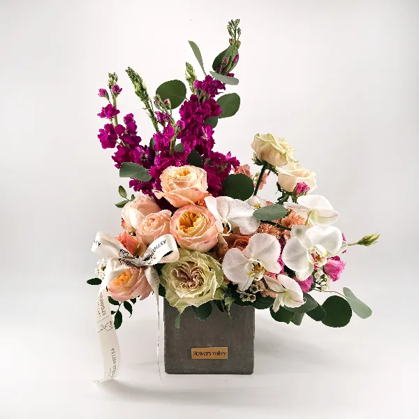 A floral arrangement featuring peach roses, white orchids, and purple stock in a grey box with a Flowers Valley label.