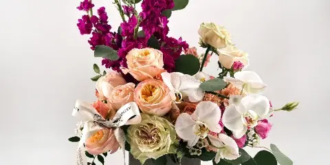 A floral arrangement featuring peach roses, white orchids, and purple stock in a grey box with a Flowers Valley label.