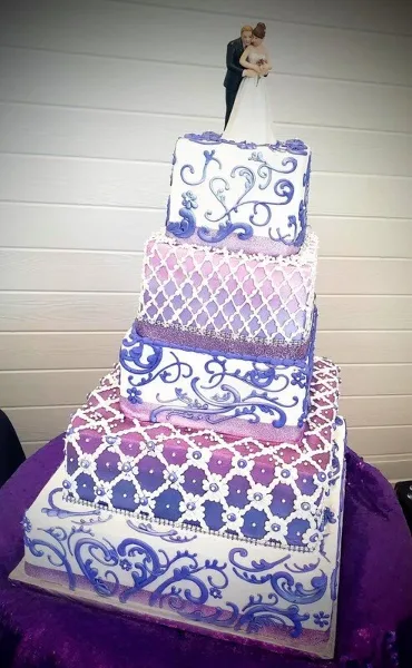 A five-tier square wedding cake decorated with purple and white patterns and a bride and groom topper.
