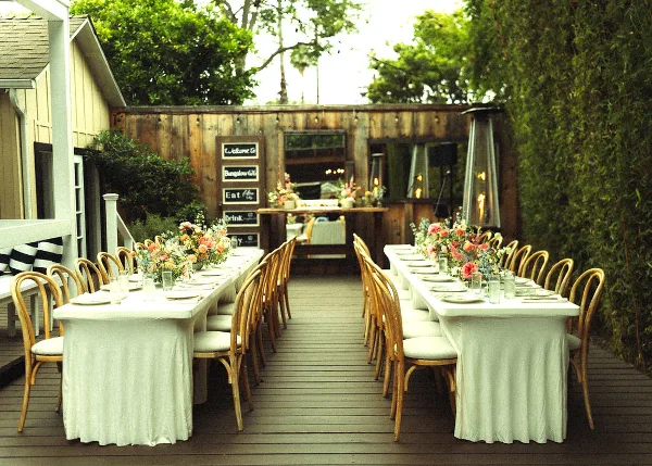 An outdoor event space featuring two long tables set with white linens, wooden chairs, and floral centerpieces, with a wooden structure in the background.
