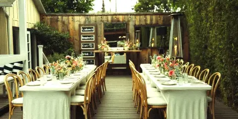 An outdoor event space featuring two long tables set with white linens, wooden chairs, and floral centerpieces, with a wooden structure in the background.