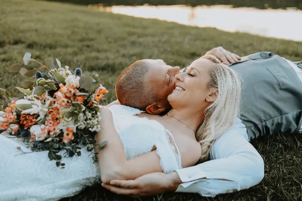 A bride and groom lying on the grass in a field, with the bride holding a floral bouquet.
