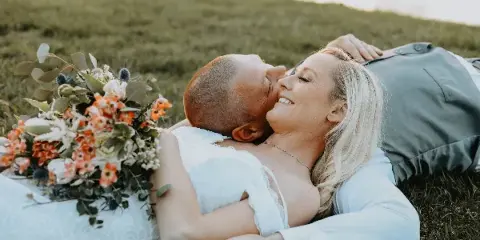 A bride and groom lying on the grass in a field, with the bride holding a floral bouquet.