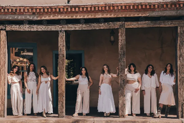 A group of eight women in white and cream wedding attire standing in front of a rustic adobe building.
