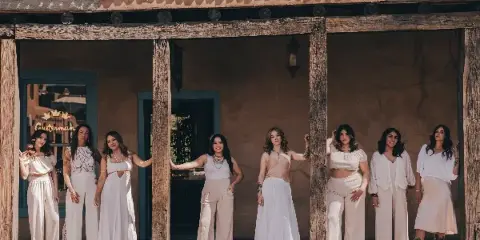 A group of eight women in white and cream wedding attire standing in front of a rustic adobe building.