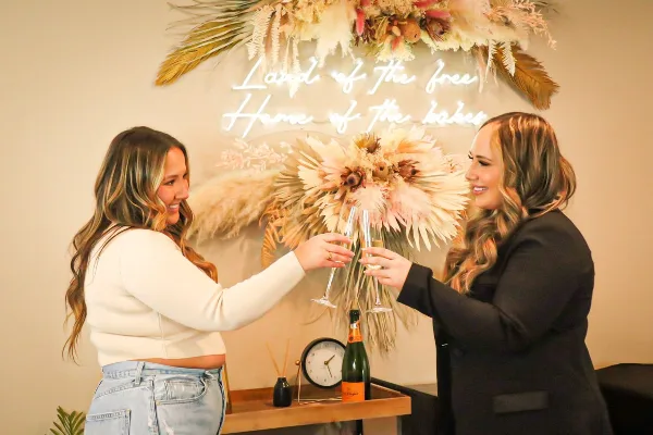 Two women smiling and toasting with champagne flutes in front of a floral and neon sign backdrop.