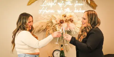 Two women smiling and toasting with champagne flutes in front of a floral and neon sign backdrop.