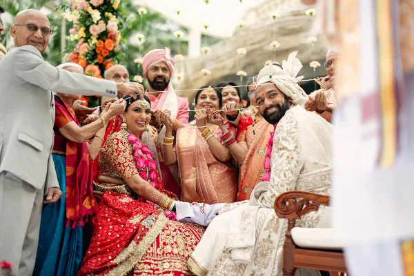 A bride and groom participate in a traditional Hindu wedding ceremony, holding a sacred thread together while surrounded by family members.