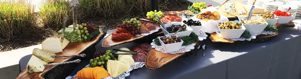 Outdoor catering table with charcuterie, cheeses, grapes, olives, and breads arranged on wooden boards and white bowls.