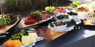 Outdoor catering table with charcuterie, cheeses, grapes, olives, and breads arranged on wooden boards and white bowls.