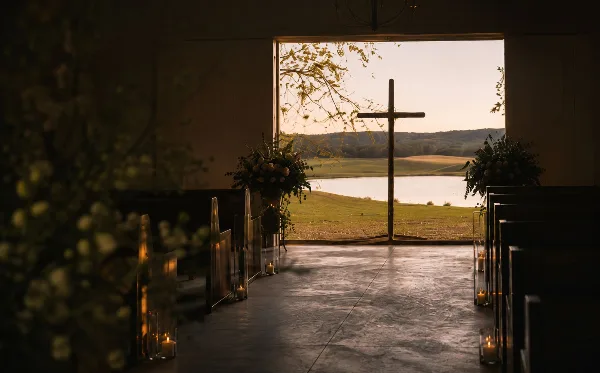 Interior view of a rustic chapel with a wooden cross, pews, and floral arrangements, looking out onto a lake and rolling hills at sunset.