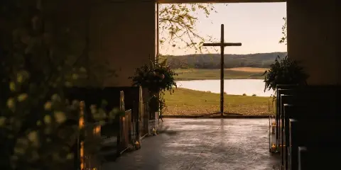 Interior view of a rustic chapel with a wooden cross, pews, and floral arrangements, looking out onto a lake and rolling hills at sunset.
