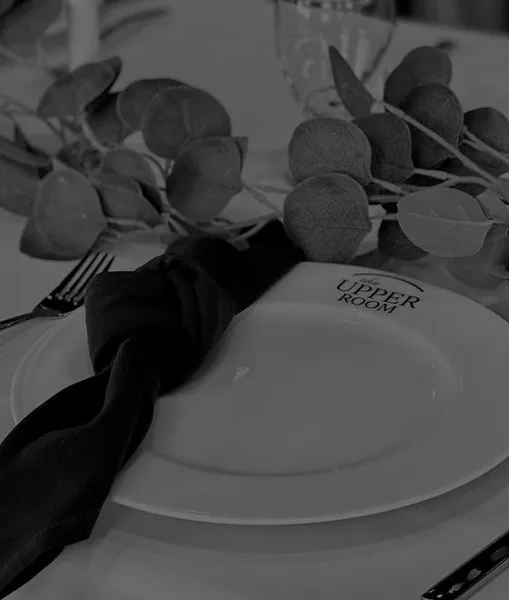 A close-up of a formal table setting featuring a white plate with 'The Upper Room' logo, a dark napkin, and eucalyptus greenery.