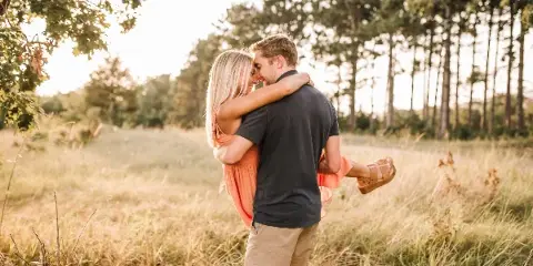 A couple embracing in a sunlit grassy field with trees in the background.