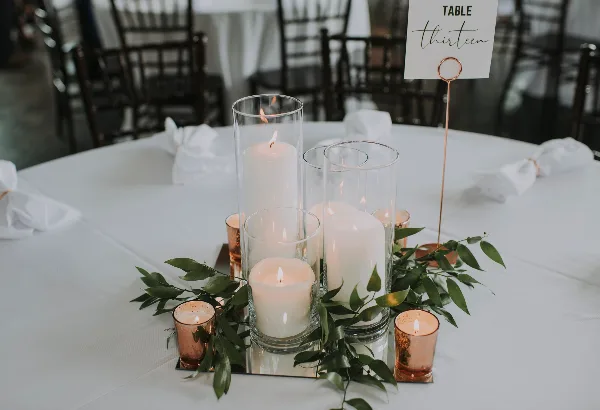 A wedding table centerpiece featuring glass cylinder vases with pillar candles and greenery on a white tablecloth.