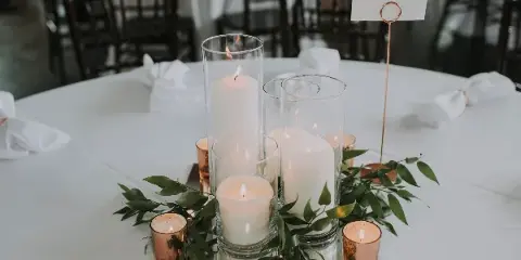 A wedding table centerpiece featuring glass cylinder vases with pillar candles and greenery on a white tablecloth.