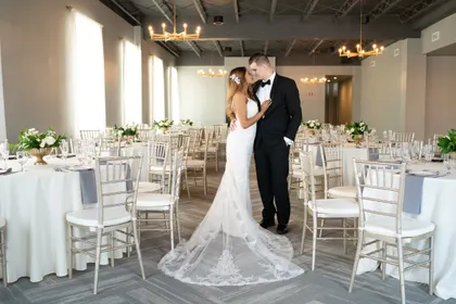 A bride and groom standing in a wedding reception hall with round tables, white chairs, and floral centerpieces.