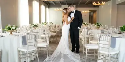 A bride and groom standing in a wedding reception hall with round tables, white chairs, and floral centerpieces.