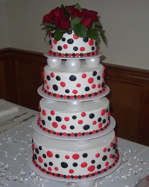 A four-tier white wedding cake decorated with red and black polka dots and topped with red roses.