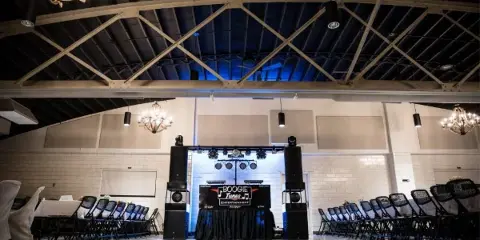 A professional DJ booth with branded table and speakers set up in a wedding reception hall.