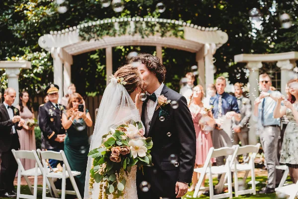 A bride and groom sharing a kiss during an outdoor wedding ceremony under a white arched structure, with guests and bubbles in the background.