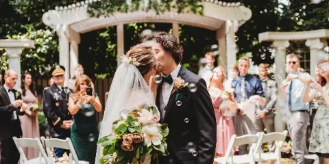 A bride and groom sharing a kiss during an outdoor wedding ceremony under a white arched structure, with guests and bubbles in the background.