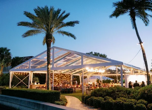 A large clear-top event tent illuminated at dusk with string lights and tables set up inside, surrounded by palm trees.
