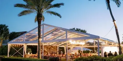 A large clear-top event tent illuminated at dusk with string lights and tables set up inside, surrounded by palm trees.
