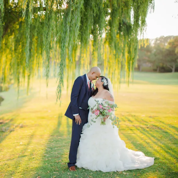 A bride and groom standing on a golf course green under a weeping willow tree.