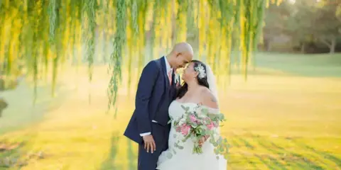 A bride and groom standing on a golf course green under a weeping willow tree.