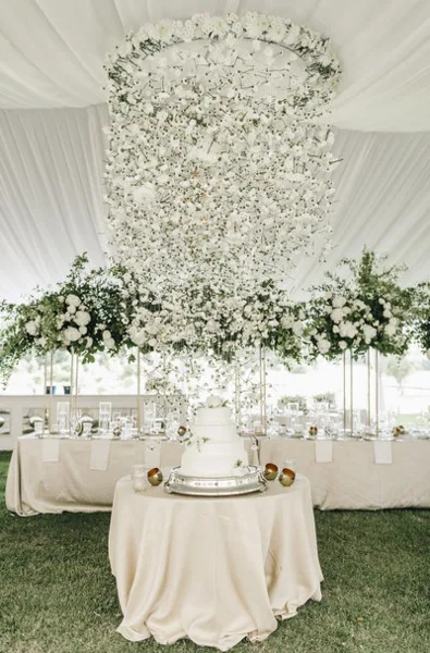 Luxury wedding reception featuring a large hanging floral chandelier and a tiered cake on a draped table.