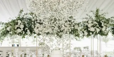 Luxury wedding reception featuring a large hanging floral chandelier and a tiered cake on a draped table.
