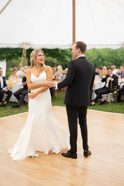 A bride and groom dancing on a wooden dance floor at an outdoor wedding reception under a white tent.