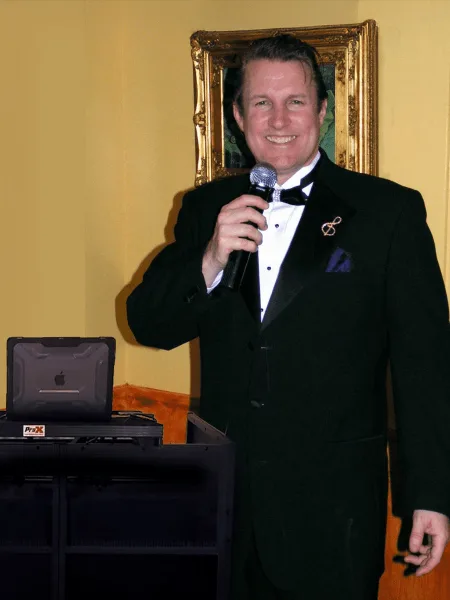 A man in a tuxedo holding a microphone standing next to a DJ laptop and equipment rack.
