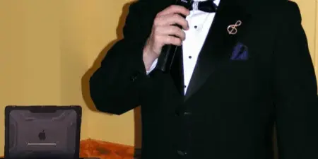 A man in a tuxedo holding a microphone standing next to a DJ laptop and equipment rack.