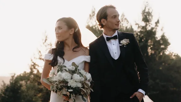 A bride in a white off-the-shoulder gown holding a bouquet and a groom in a black tuxedo standing together outdoors.