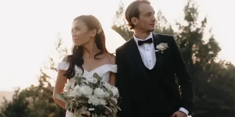 A bride in a white off-the-shoulder gown holding a bouquet and a groom in a black tuxedo standing together outdoors.