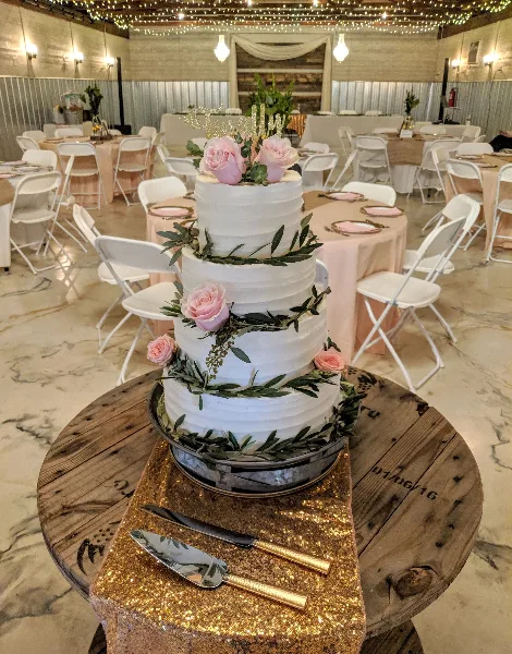 A four-tier white wedding cake decorated with pink roses and greenery, displayed on a wooden stand in a banquet hall.
