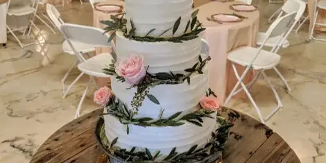 A four-tier white wedding cake decorated with pink roses and greenery, displayed on a wooden stand in a banquet hall.