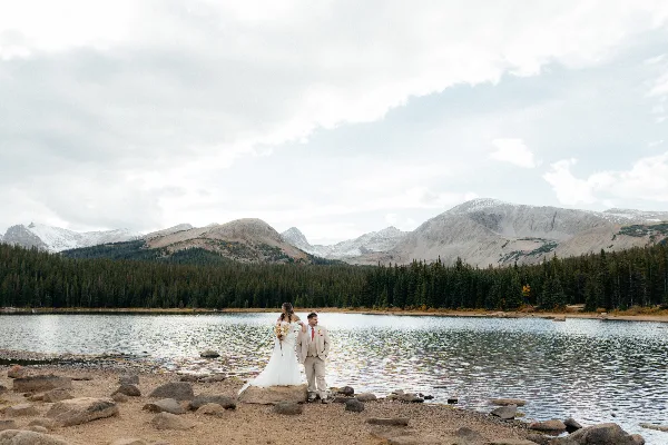 A bride and groom standing on a rocky shore by a mountain lake with snow-capped peaks in the background.