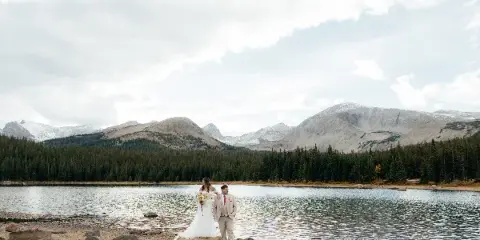 A bride and groom standing on a rocky shore by a mountain lake with snow-capped peaks in the background.