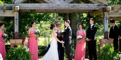 Ann McKenzie officiating a wedding ceremony outdoors with a bride and groom at an altar, surrounded by bridesmaids and groomsmen.