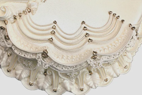 Close-up photograph of an ornate white wedding cake with detailed piping and gold accents.