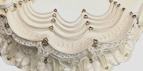 Close-up photograph of an ornate white wedding cake with detailed piping and gold accents.