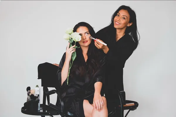 A makeup artist applying makeup to a seated client using a brush, with makeup tools visible on a nearby table.