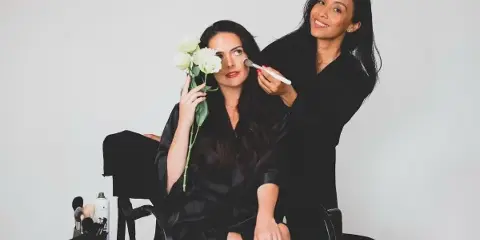 A makeup artist applying makeup to a seated client using a brush, with makeup tools visible on a nearby table.