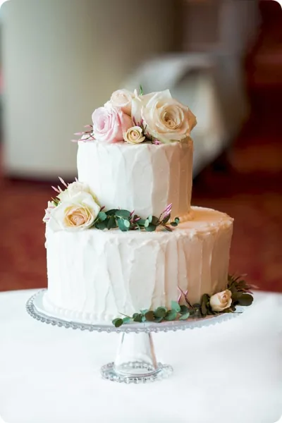 Two-tier white frosted wedding cake topped with fresh pink and cream roses and eucalyptus, displayed on a glass cake stand.