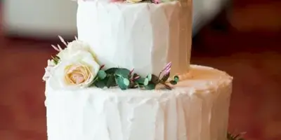 Two-tier white frosted wedding cake topped with fresh pink and cream roses and eucalyptus, displayed on a glass cake stand.