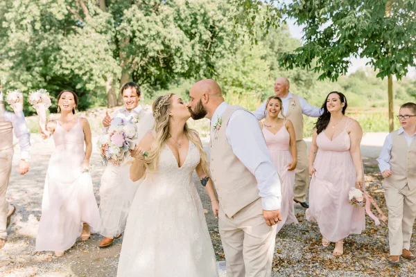 A bride and groom share a kiss during their wedding, surrounded by their smiling wedding party in an outdoor setting.