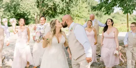 A bride and groom share a kiss during their wedding, surrounded by their smiling wedding party in an outdoor setting.
